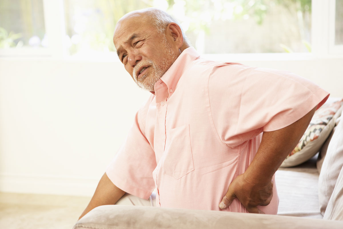 An older man sitting in a chair touching his lower back.
