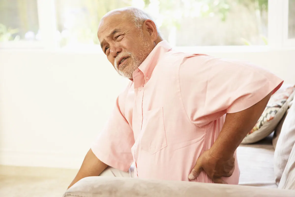 An older man sitting in a chair touching his lower back.