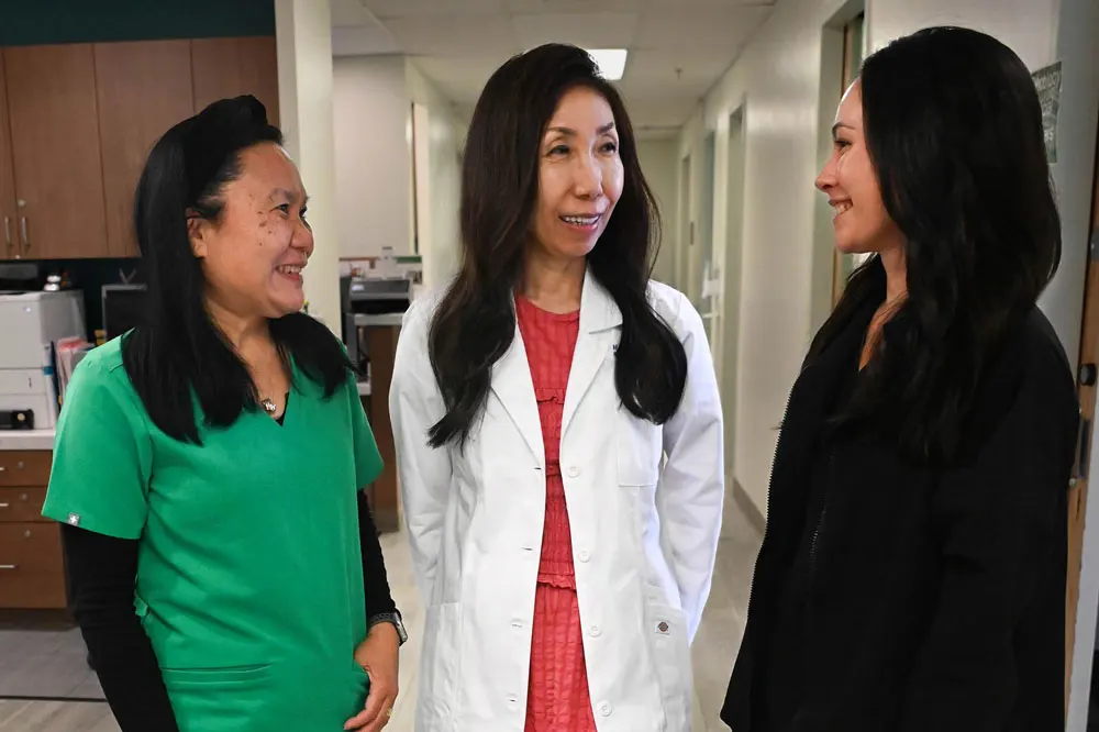 Doctor in white coat beside two colleagues, an MA in green scrubs and a pharmacist in black.