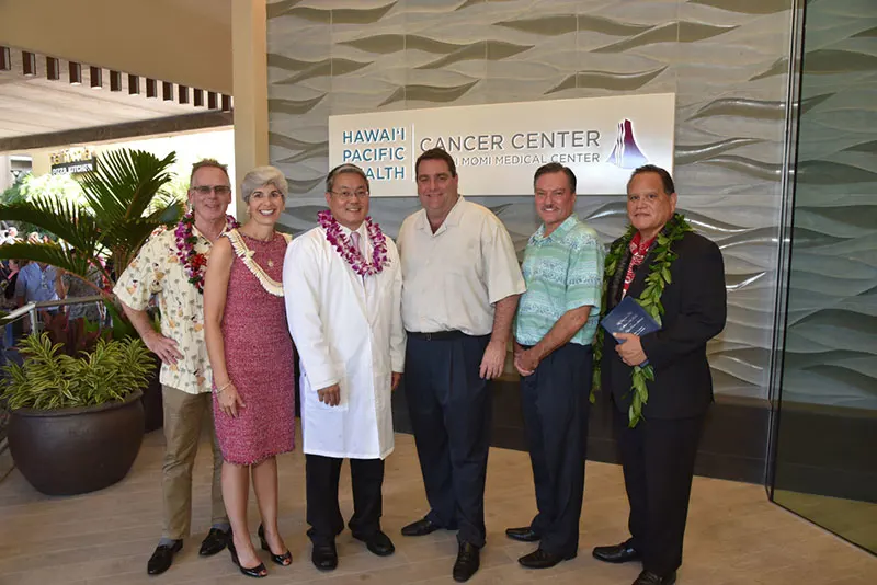 Group shot of executives and stakeholders wearing lei in front of sign for the Hawaii Pacific Health Cancer Center at Pali Momi Medical Center.