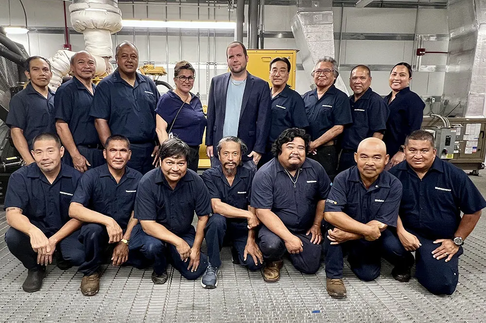Large group with two rows of facilities staff in black polo shirts in a utility room at Wilcox Medical Center.