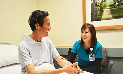 Nurse in chair and a patient sitting up in bed laugh together.