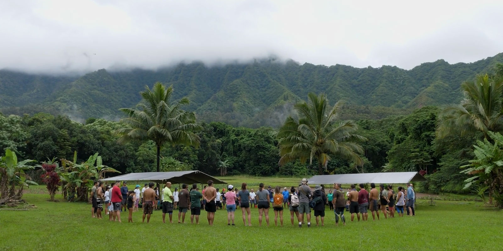 Circle of people in a vibrant green outdoor area with green mountains in the background and dramatic low hanging clouds.