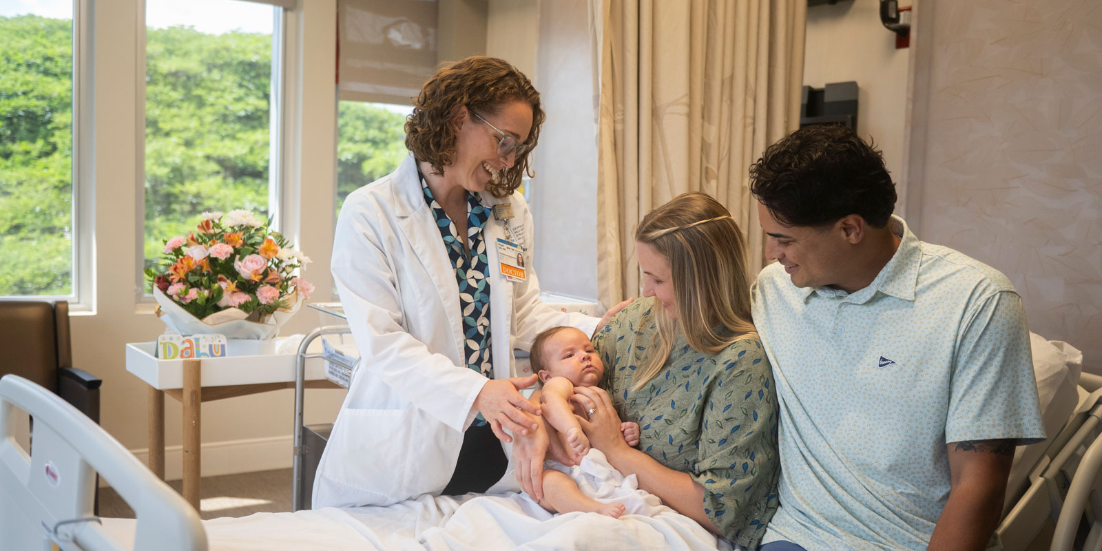 a doctor stands and smiles at a mother, father, and their newborn baby as they sit on a bed in a light-filled room in the Mother Baby Care Unit at Kapiolani Medical Center for Women & Children