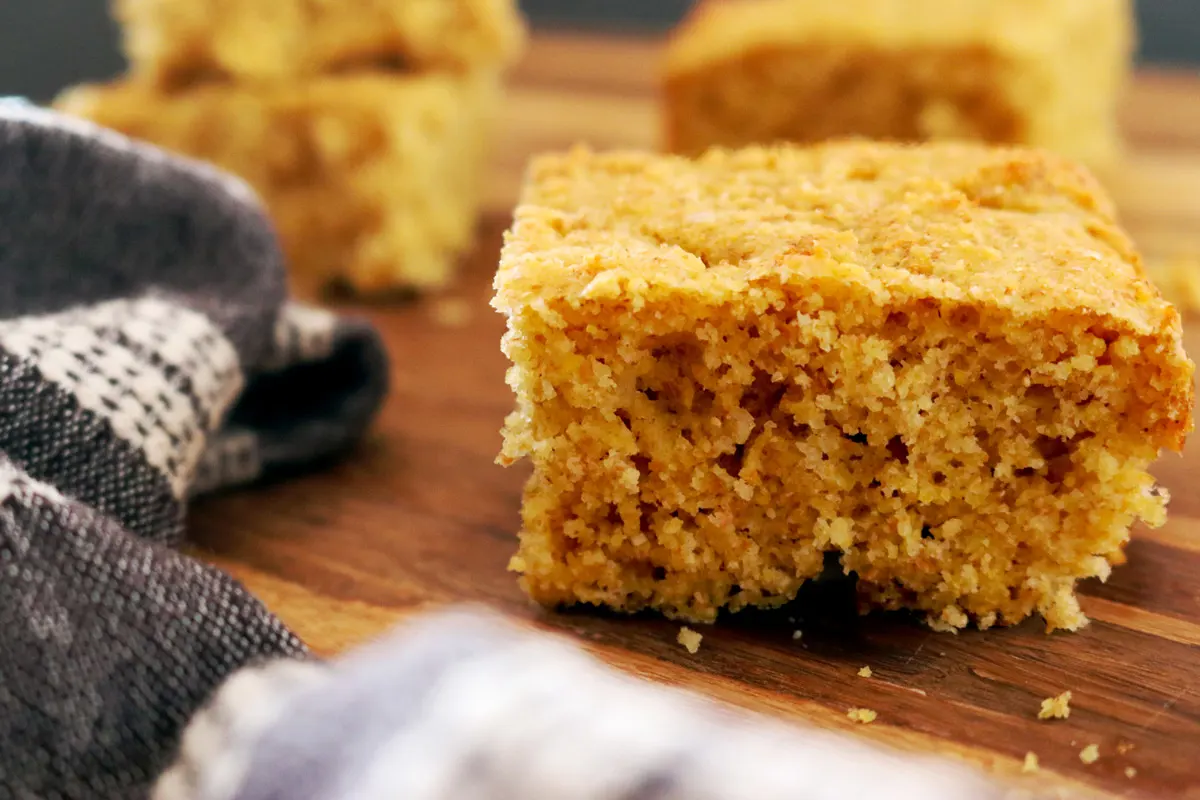 A closeup of cornbread slices on a wooden butcher block counter with a dark-grey hand towel placed off to the side.