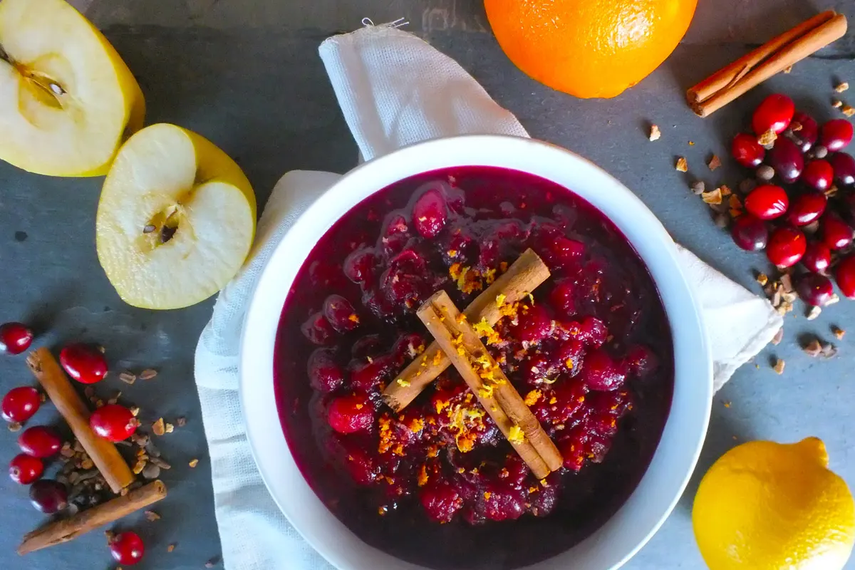 Bright cranberry chutney, garnished with cinnamon sticks and citrus zest sits in a white bowl.