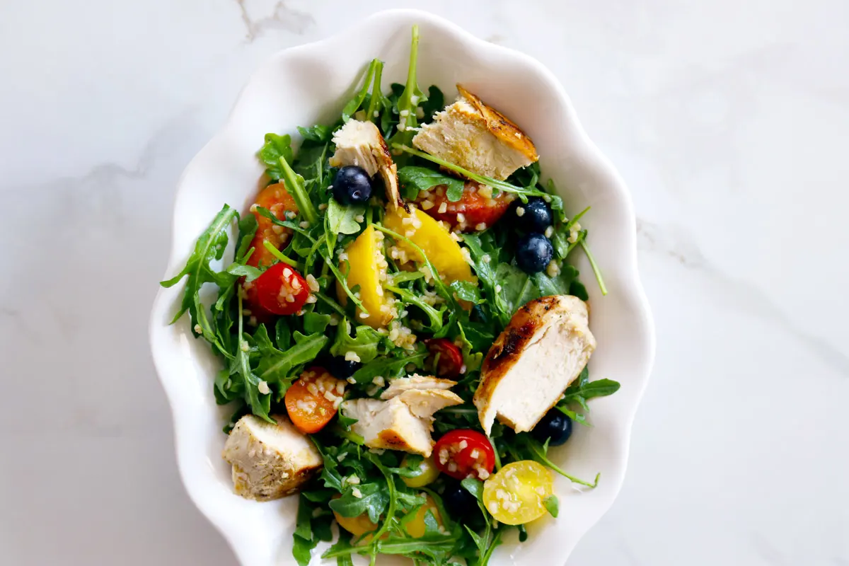 A white bowl filled with Chicken & Bulgur Salad made with arugula, grilled chicken, bulgur, cherry tomatoes, peaches and blueberries sits on a white marble counter top.