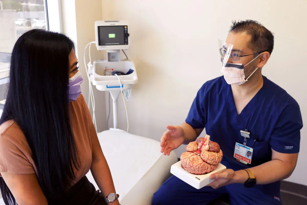 A male doctor holds a model of a brain and talks with a female patient as they sit in an exam room.
