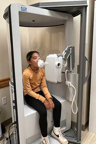 Child sitting inside a special booth, breathing into equipment to test lung function.