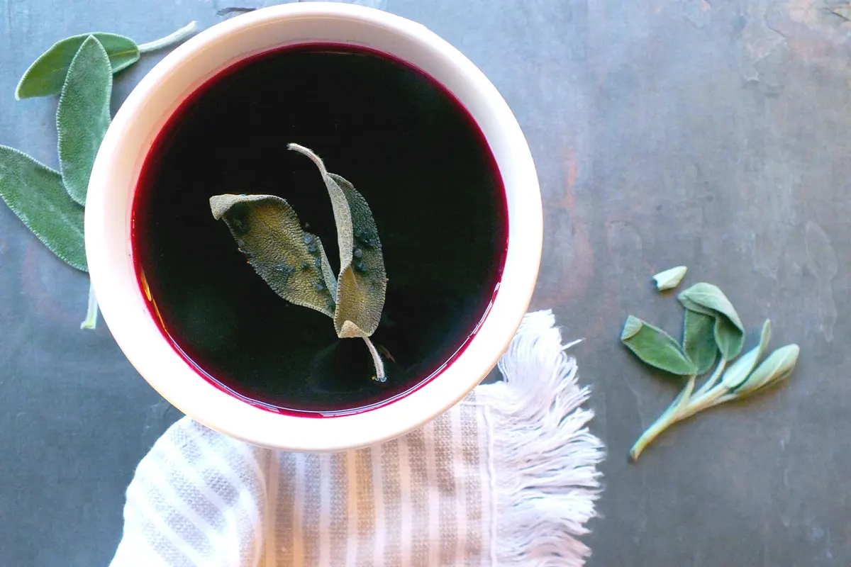 Deep red beet soup is pictured in a white bowl with sage garnish.